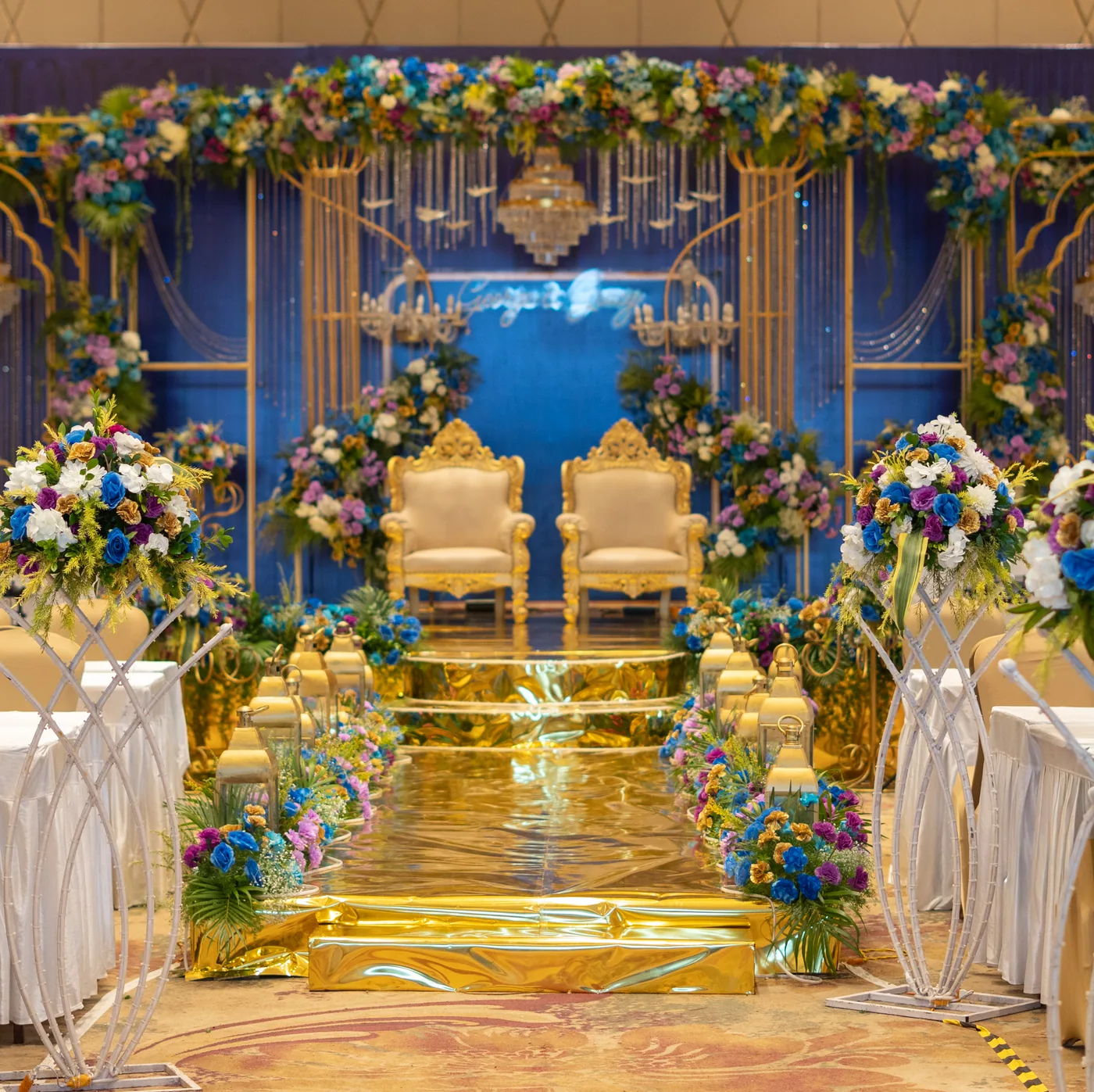 Reception stage in royal blue and gold, with carved gold chairs, floral arches and a gold mirrored aisle