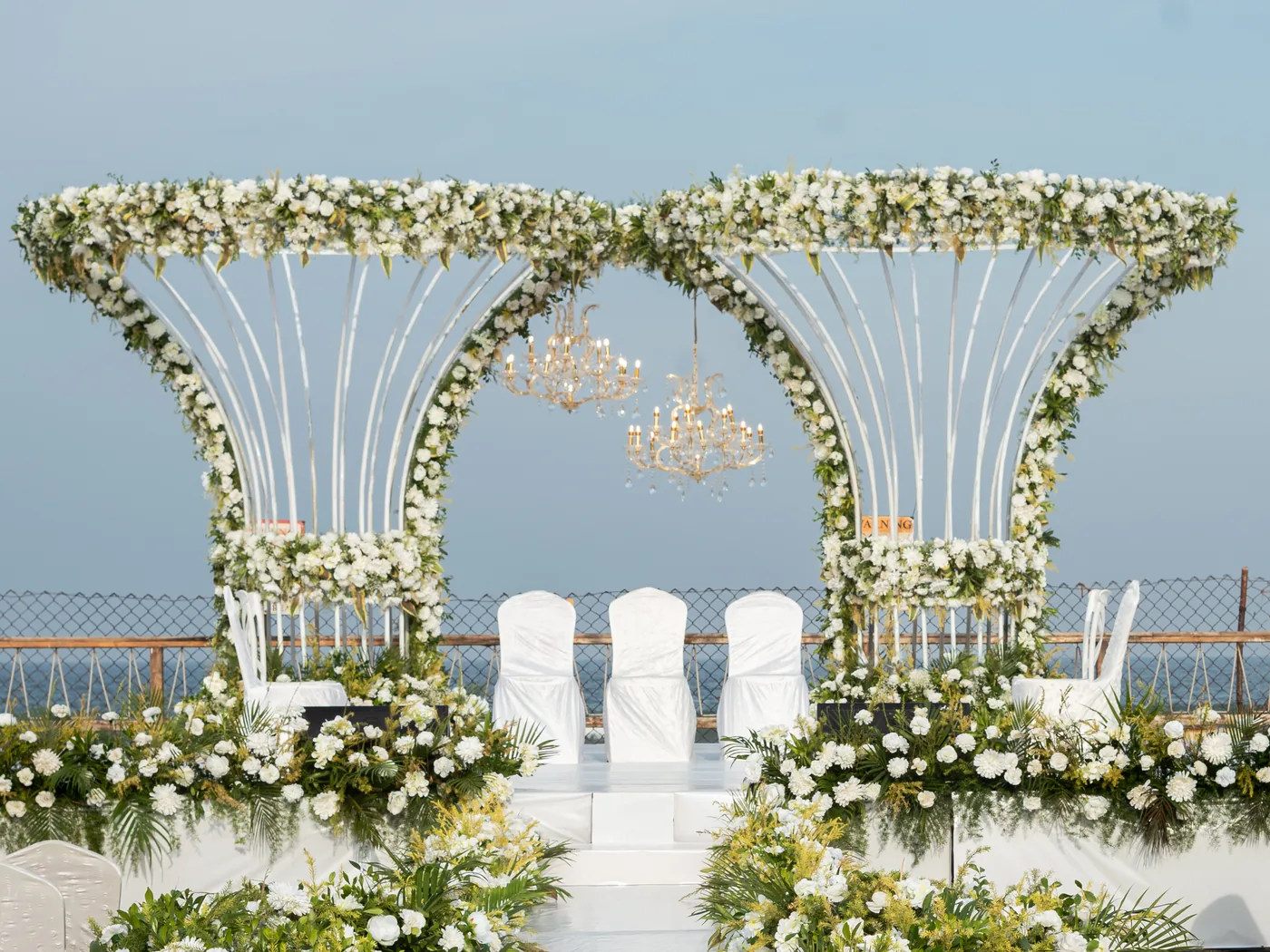Front view of a white floral twin-arch wedding stage with chandeliers, chairs and the sea behind