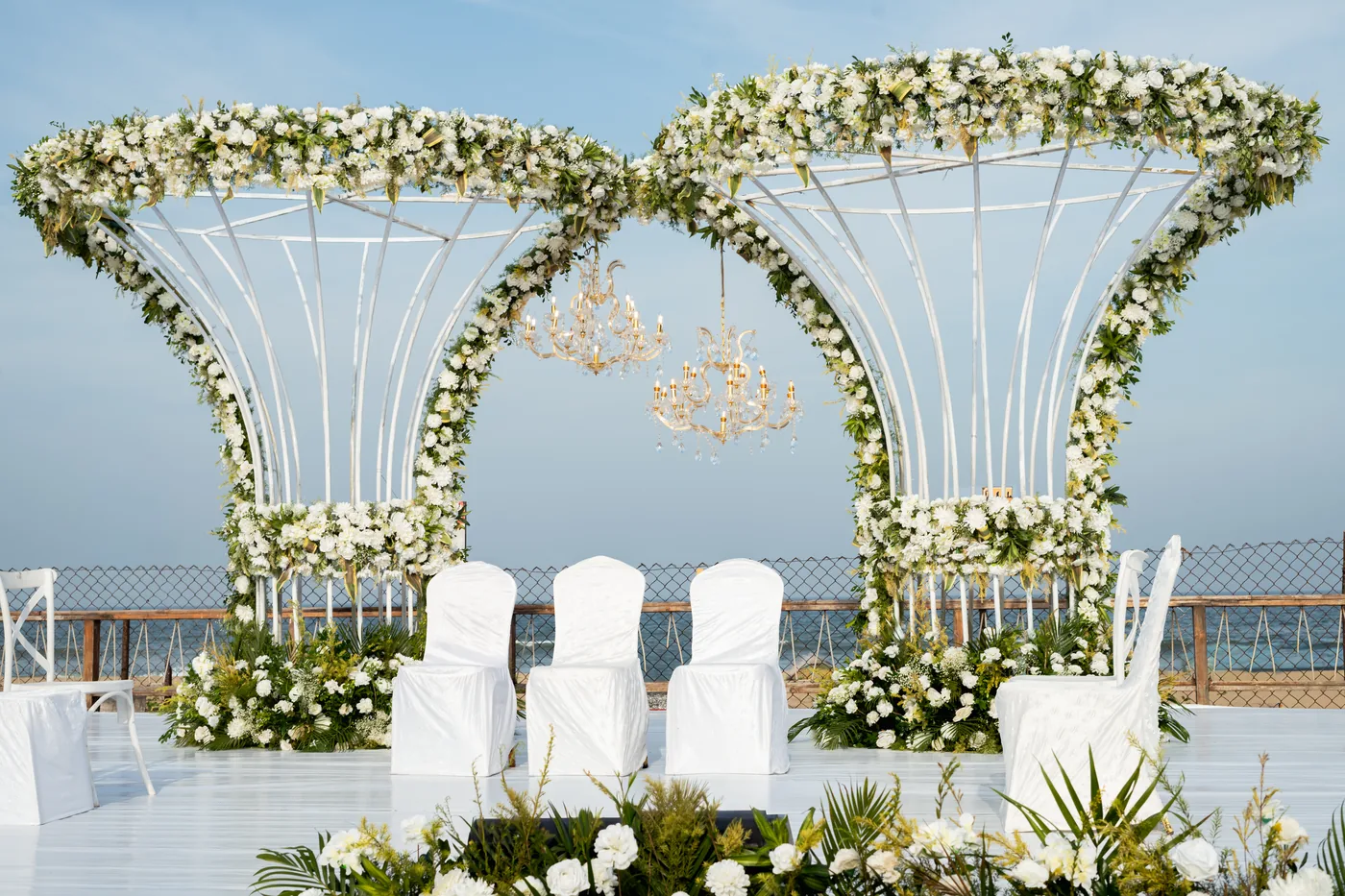 Twin flared arches of white flowers with crystal chandeliers hanging inside, on a white wedding stage