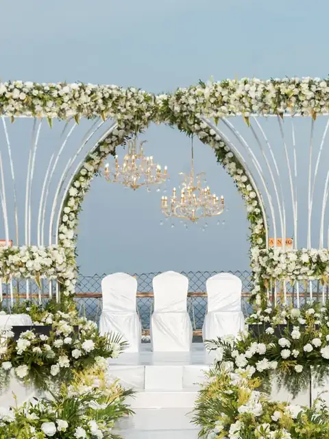 Front view of a white floral twin-arch wedding stage with chandeliers, chairs and the sea behind